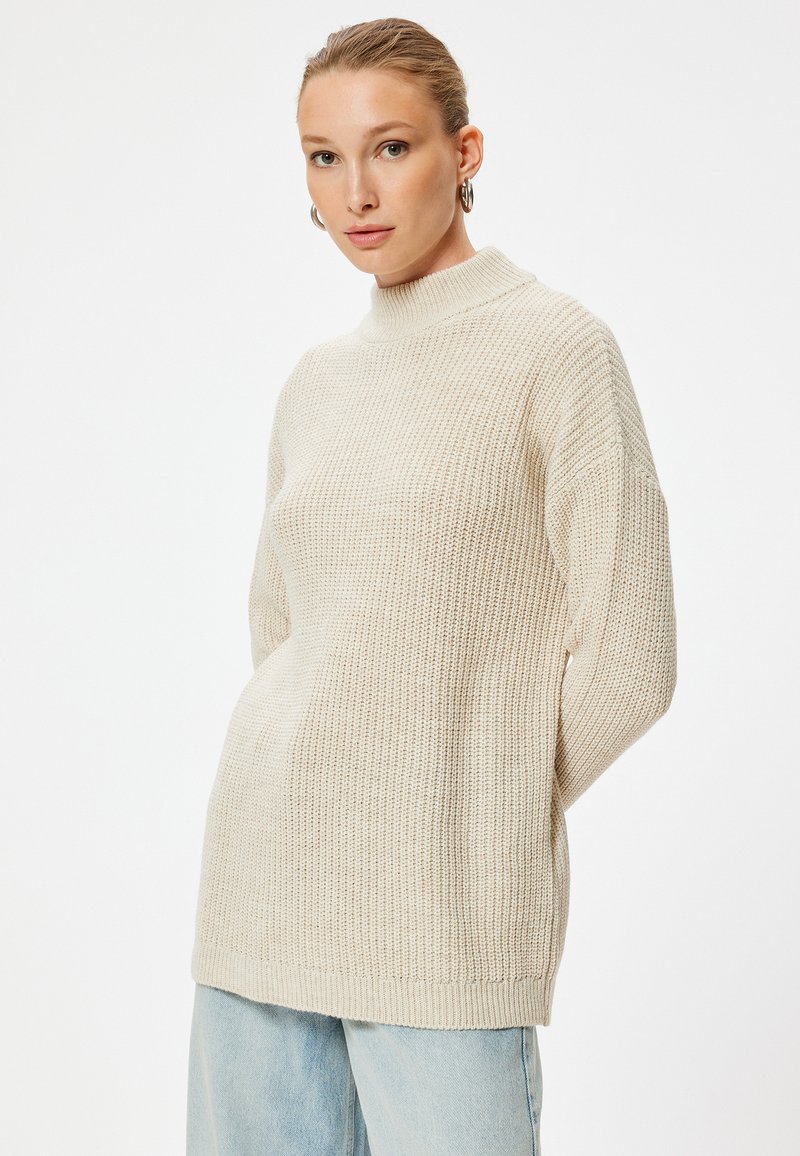 Cremig gestrickter Pullover mit hohem Kragen, lockerer Passform und strukturierter Oberfläche. Kombiniert mit hellblauer Jeans.