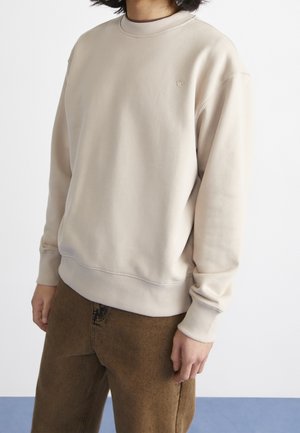 Personne portant un sweat-shirt beige à col rond avec un petit logo brodé sur la poitrine et un pantalon marron, debout devant un fond uni.