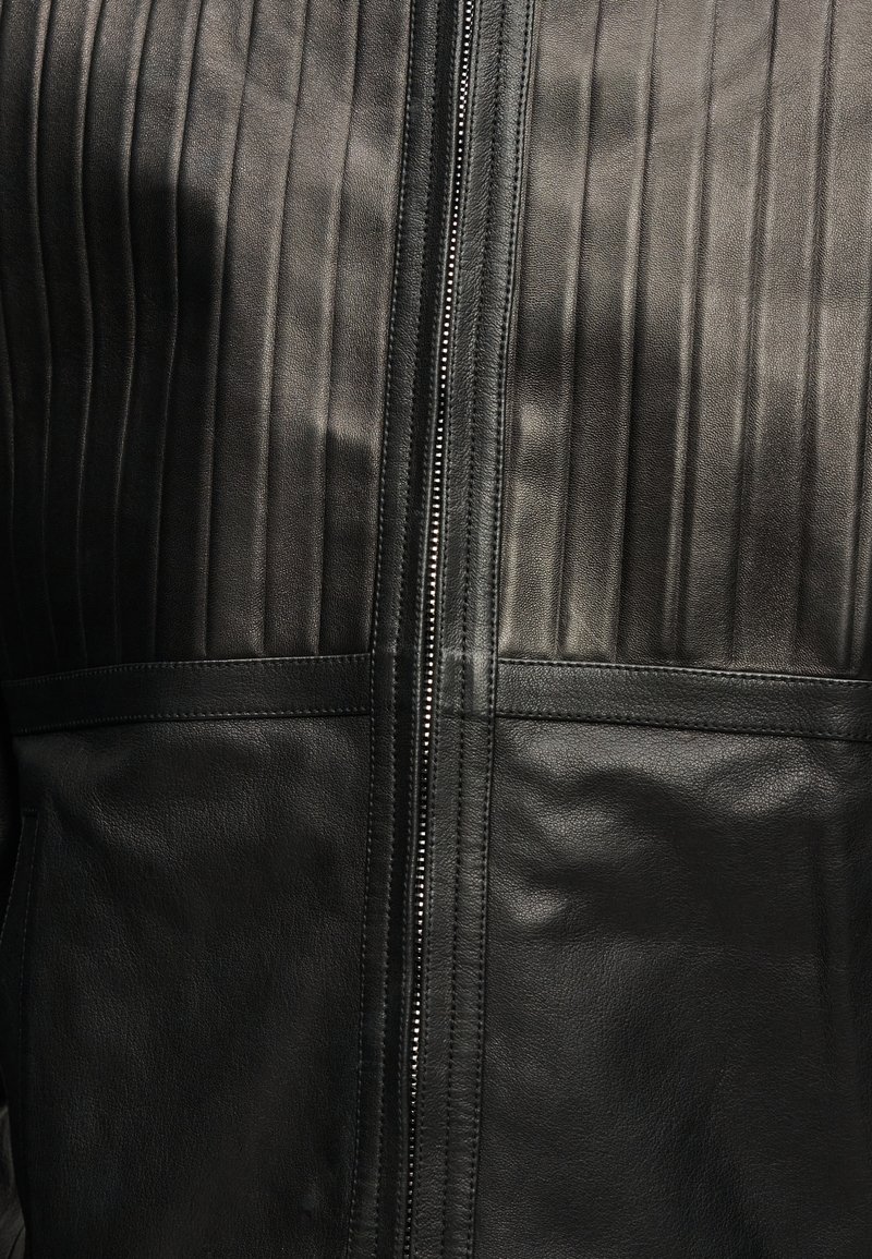 Veste en cuir noir avec des panneaux verticaux cousus en haut et du cuir lisse en bas, équipée d'une fermeture éclair argentée centrée.