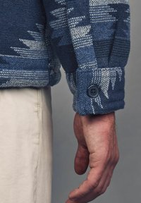 Texturovaná modrá tkaná bunda s vzorovaným designem a detailem na manžetách s knoflíky, zobrazená vedle ruky, na neutrálním pozadí.