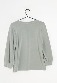 Lichtgrijs langarm T-shirt met elastische manchetten hangt aan een zwarte hanger tegen een witte achtergrond.