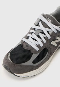 Grå atletisk sko med ovandel i mocka och mesh. Har vita snörningar, logotypdetalj och en texturerad gummisula. Närbild av ovandelen.