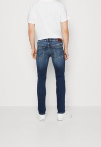 Man som bär slim-fit mörkblå jeans med bakfickor och vita sneakers, stående mot en enkel vit bakgrund.