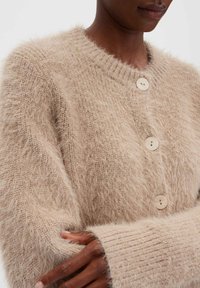Cardigan beige en peluche avec un col rond, quatre grands boutons et des poignets côtelés, doté d'une finition douce et texturée.
