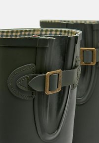 Ciemnozielone gumowe kalosze z podszewką w kratkę. Wśród cech znajdują się dekoracyjne klamry oraz mosiężne akcenty po boku.