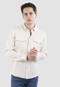 Lys beige skjorte med knapper, to brystlommer, klassisk krave, lange ærmer og buet kant, parret med blå denim jeans.