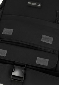 Mochila de tela negra con una cremallera prominente, parches de Velcro y un hebilla de plástico para el cierre. Diseño minimalista con un layout organizado.