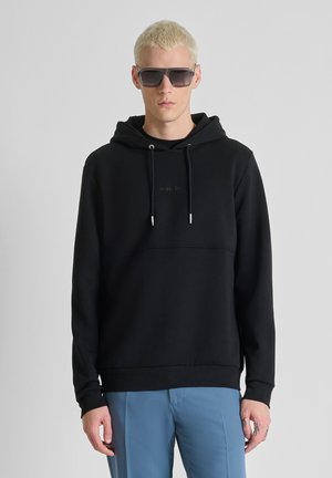 Joven con cabello rubio corto, vestido con sudadera con capucha negra, gafas de sol grandes y pantalones azules, de pie frente a un fondo gris liso.