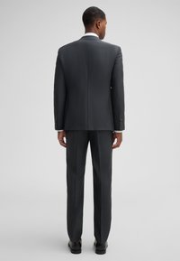 Dunkelgrauer, maßgeschneiderter Anzug mit einem Kerbkragen, einem einfachen Schlitz und flachen Hosen. Strukturierter Stoff mit dezenten Mustern. Schwarze Schuhe.