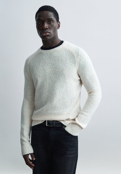 Cream textured sweater with ribbed cuffs, round neckline, and contrasting black inner collar. Paired with dark denim jeans and a black belt.