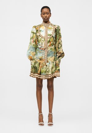 Moteris stovinti, dėvinti ilgomis rankovėmis, aukštu kaklu su tropikų raštu, siaurintu liemeniu, dryžuotu apačia ir baltais kulnais su čiurnos dirželiais ant balto fono.