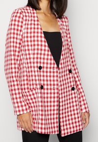Blazer s kostkovaným vzorem v červené a bílé barvě, s V výstřihem, dvouřadým zapínáním na černé knoflíky a dlouhými rukávy. Nositelný přes černý top.