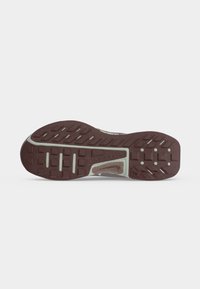 Semelle d'une chaussure de course Nike Trail marron et grise avec des motifs de crampons rectangulaires et circulaires sur fond blanc.
