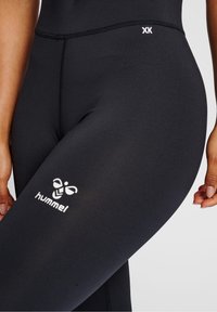 Czarne leginsy wykonane z elastycznego materiału, z białym logo "hummel" i małą metką "XK" w pobliżu pasa. Gładka faktura.