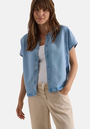 Frau mit langen braunen Haaren, die ein hellblaues Kurzarmshirt, ein weißes Top und eine beigefarbene Hose trägt, steht mit einer Hand in der Tasche.