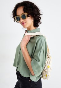 Camisa verde de manga curta, jeans pretos e uma bolsa de croché creme com detalhes em flores amarelas. Óculos de sol transparentes oversized.