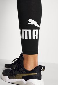 Czarne legginsy sportowe z białym logo Puma i sylwetką kota. W zestawie czarne buty sportowe z złotym akcentem i białą podeszwą.