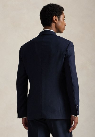Man wearing a dark navy suit with a white shirt, shown from the back facing slightly to the right against a plain background.
