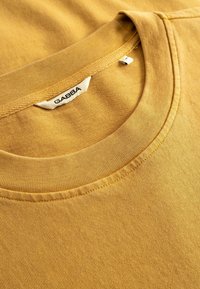 Κοντινό πλάνο ενός μουσταρδί κίτρινου T-shirt με στρογγυλή λαιμόκοψη που δείχνει την ετικέτα της μάρκας GABBA ραμμένη μέσα στο γιακά.