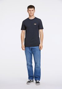 Camiseta de algodón azul marino de manga corta, con cuello redondo, que presenta un pequeño logo multicolor en el lado izquierdo del pecho, combinada con vaqueros azules y zapatillas.