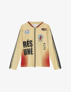 Béžová bunda na zips s dlhým rukávom s červeným prechodom, s čiernym grafickým textom a viacerými logami na prednej strane a rukávoch, vyrobená z ľahkej tkaniny.