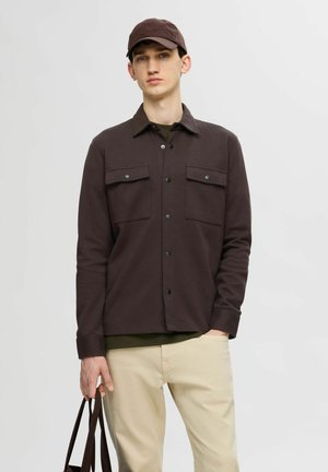 Chemise brune foncée à boutons avec deux poches poitrine, associée à une casquette noire et un pantalon beige clair. Le tissu semble lisse et structuré.
