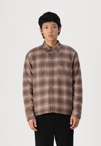 Camicia a quadri marrone con bottoni, realizzata in tessuto morbido, dotata di colletto, due tasche sul petto e una vestibilità leggermente rilassata.