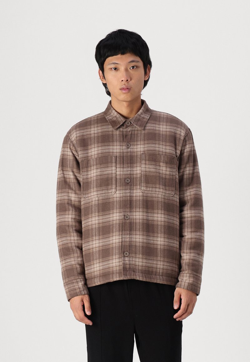 Camicia a quadri marrone con bottoni, realizzata in tessuto morbido, dotata di colletto, due tasche sul petto e una vestibilità leggermente rilassata.
