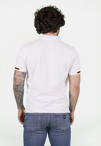 Polo shirt bianco a maniche corte con colletto e maniche impreziosite da righe nere, rosse e gialle. Indossato con jeans blu.