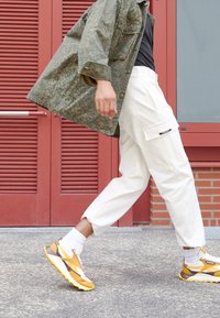 Modell iklädd en mönstrad olivgrön jacka, svart skjorta, vita cargobyxor med sidofickor och sneakers med gula accenter, går.