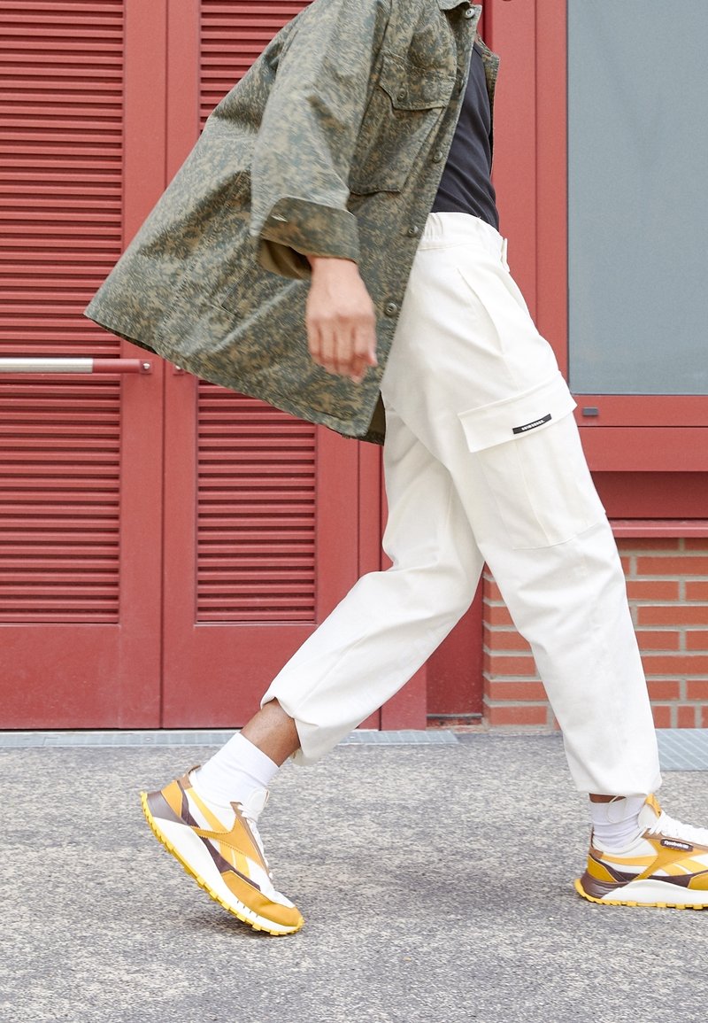 Modell iklädd en mönstrad olivgrön jacka, svart skjorta, vita cargobyxor med sidofickor och sneakers med gula accenter, går.