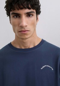 Camiseta de algodón azul marino con cuello redondo, que presenta el texto bordado en blanco "Scalpers Company" en el lado izquierdo del pecho. Textura suave.