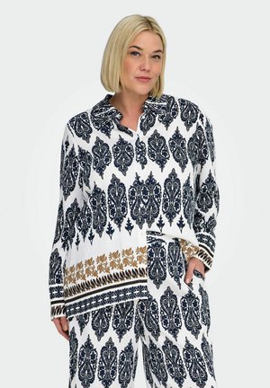 Žena s krátkymi blond vlasmi oblečená v bielom outfite so zložitými modrými a hnedými kvetinovými a geometrickými vzormi, pózujúca s jednou rukou v vrecku.