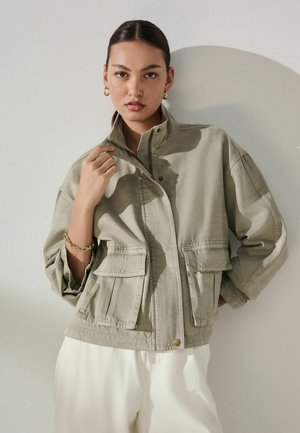 Frau mit zurückgekämmtem Haar trägt eine hellgrüne, übergroße Jacke mit Patttaschen und weiße Hose, posiert vor einem schlichten weißen Hintergrund.