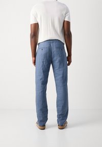 Lichtblauwe linnen broek met een relaxte pasvorm, voorzien van twee achterzakken en een elastische tailleband met trekkoord. G gedragen met een wit shirt en sandaal.