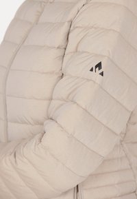 Ljusbeige pufferjacka med quiltat mönster, med en svart logotyp på vänster ärm och en slät, mjuk textur.