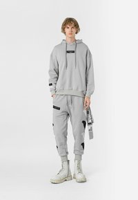 Sudadera gris claro y pantalones de chándal a juego con acentos negros. Capucha con cordones, tejido texturizado y detalles gráficos en los pantalones. Botas blancas.
