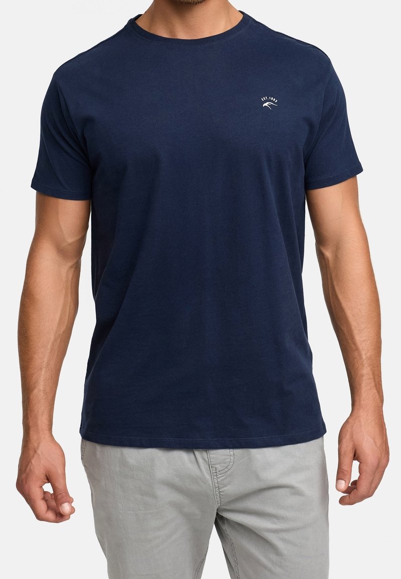 Homme portant un t-shirt bleu marine uni à manches courtes avec un petit logo blanc sur la poitrine gauche et un pantalon gris clair.