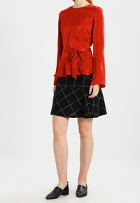 Top rosso a maniche lunghe con vita a peplum e dettaglio con fiocco, abbinato a una gonna nera con un motivo a griglia bianca. Scarpe nere flat completano l'outfit.