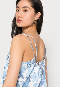 Camisola fluida azul com padrão floral, com alças de espaguete ajustáveis, detalhe em nó nas costas e textura de tecido suave.