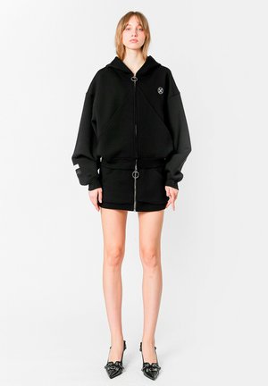 Joven mujer que lleva una sudadera con capucha negra de gran tamaño con cremallera, una mini falda y tacones negros de punta con adornos, de pie frente a un fondo blanco.