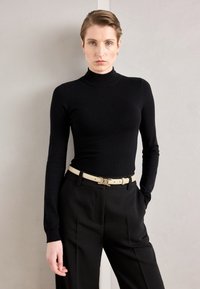 Top aderente nero a collo alto abbinato a pantaloni neri a vita alta. Una cintura crema con fibbia metallica valorizza l'outfit. Texture liscia.