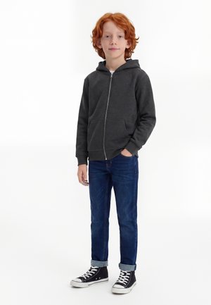 Giovane ragazzo rosso di capelli in piedi con una mano in tasca, che indossa una felpa con cappuccio grigio scuro con zip, jeans blu con risvolti e sneakers alte nere.