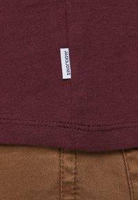 Burgunder T-Shirt aus Baumwolle mit einem kleinen weißen Markenetikett, das am Saum angenäht ist. Glatte Textur, einfaches Design, gerade Passform.