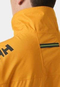 Osoba nosící žlutou bundu s vyvýšeným límcem, s černým a neonově zeleným reflexním pruhem na zádech a částečně černým logem na rameni.