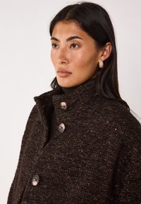 Cappotto marrone strutturato con colletto alto, dotato di grandi bottoni scuri e un motivo tessuto. Il tessuto appare spesso e caldo.