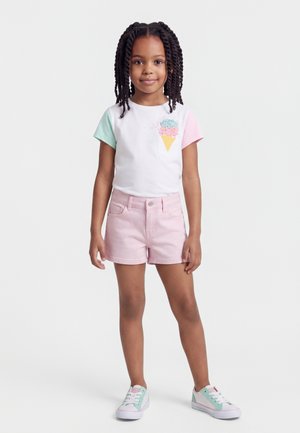 Mladé dievča s vrkočmi, oblečené v bielom tričku s motívom zmrzliny, ružových šortkách a pastelových teniskách, stojace pred jednoduchým pozadím.