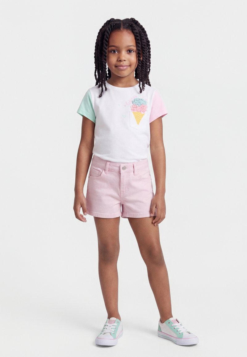 Νεαρό κορίτσι με κοτσίδες, φοράει λευκό t-shirt με σχέδιο παγωτού, ροζ σορτς και παστέλ αθλητικά παπούτσια, στέκεται μπροστά σε απλό φόντο.