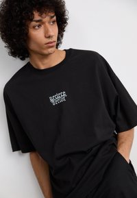 Sort oversized t-shirt i bomuld med hvid broderet tekst på brystet. Stoffet er glat, har en afslappet pasform og korte ærmer.