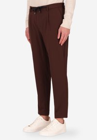 Pantaloni marroni su misura con una texture liscia, dotati di vita con cordoncino e pieghe frontali, abbinati a sneakers bianche.
